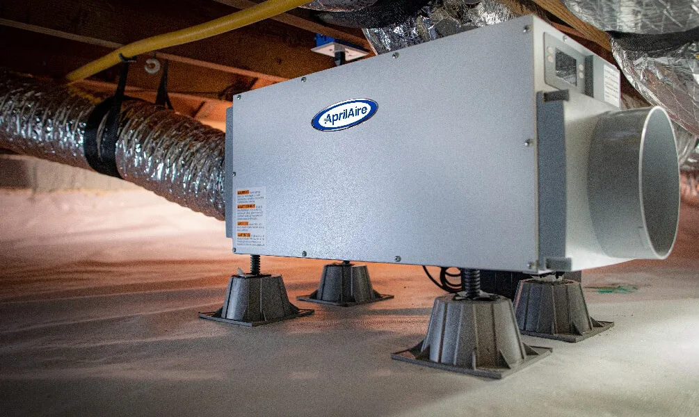 AprilAire dehumidifier unit mounted on riser feet in a white vapor barrier lined crawl space with flexible ducting.