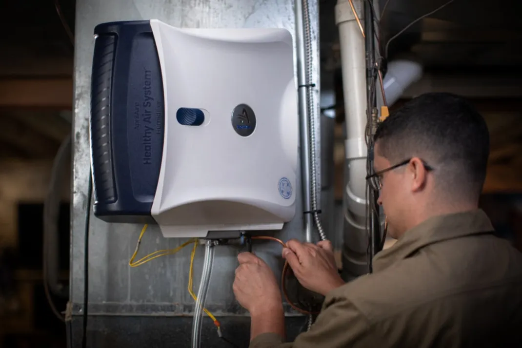 A technician installs a white AprilAire whole-home humidifier onto a metal HVAC furnace duct in a basement.