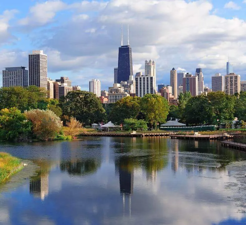 The John Hancock Center and Chicago skyscrapers reflect in a calm pond surrounded by lush green trees and a wooden boardwalk.