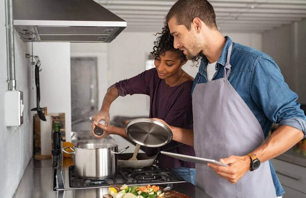 A man in an apron holds a tablet while a woman seasons a pot on a gas stove in a bright kitchen.