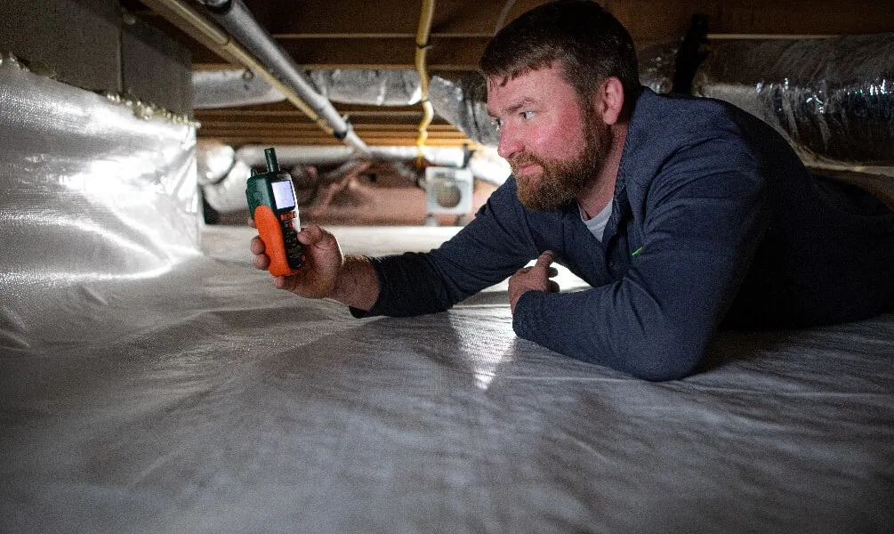 A technician crawls under a house to check moisture levels using a digital meter on a white vapor barrier.