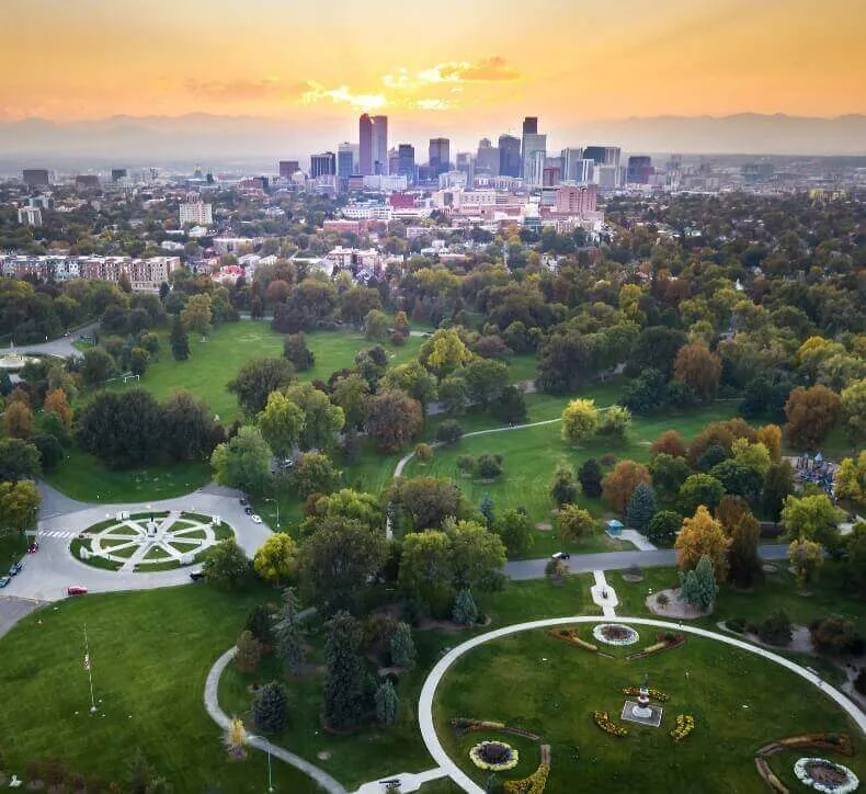 Aerial view of Denver's skyline and City Park during a golden sunset with the Rocky Mountains in the background.