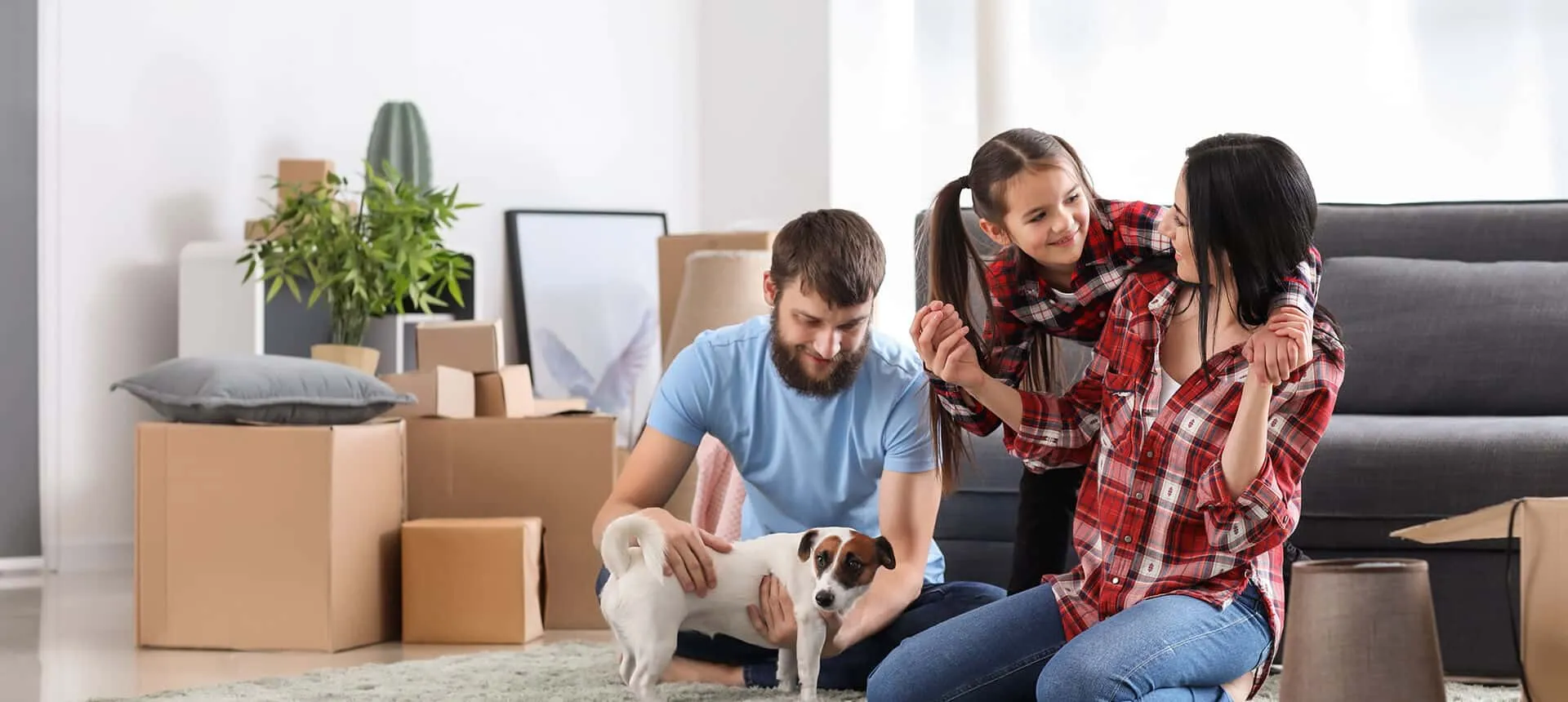 A mother, father, and young daughter sit on a living room rug with their dog surrounded by cardboard moving boxes.