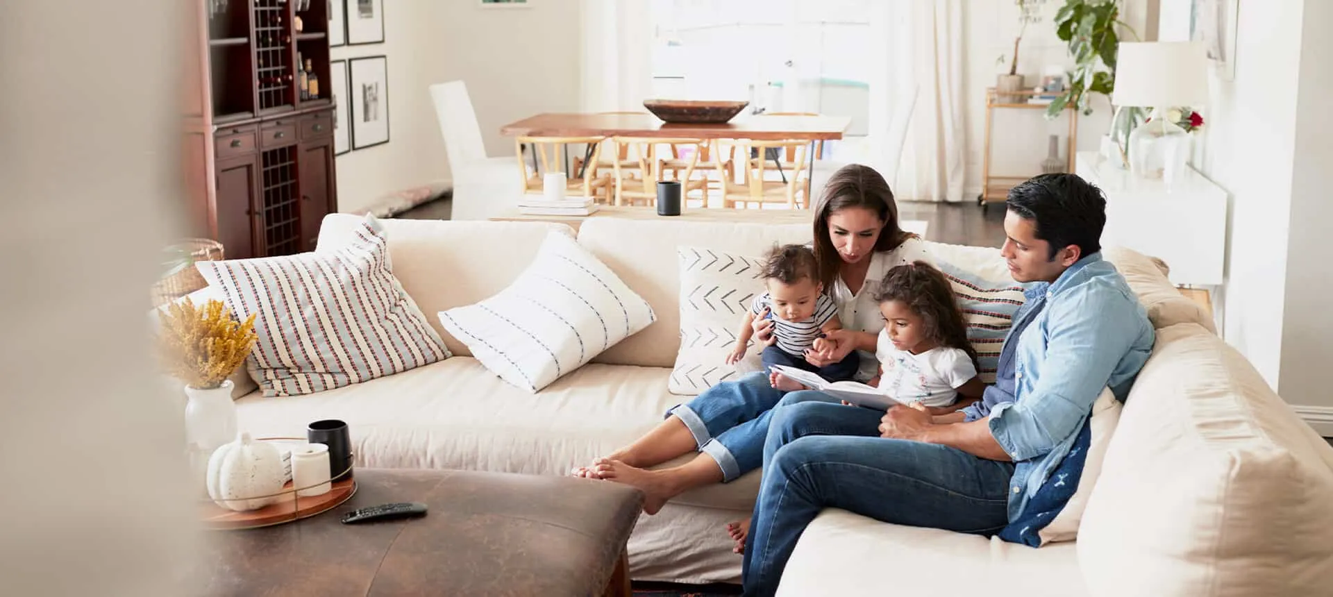 A young family with two small children sits together on a large beige sectional sofa in a bright, modern living room.