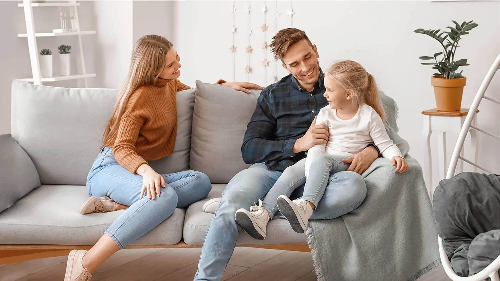 A smiling mother, father, and young daughter sitting together on a grey couch in a bright, modern living room.