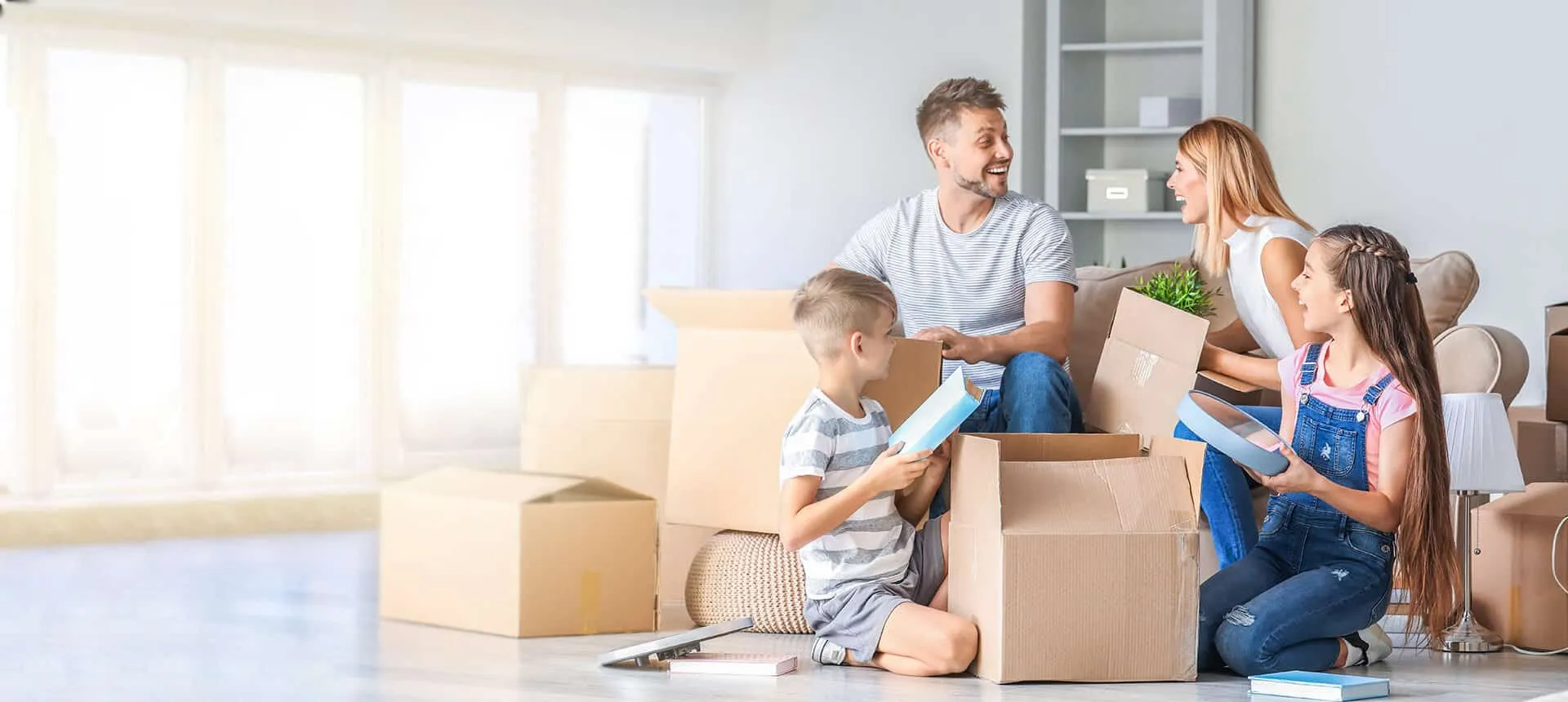 A smiling family of four sits on the floor of a bright living room unpacking cardboard boxes together.