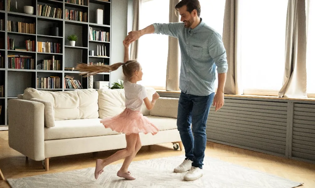 A smiling father spins his young daughter, who wears a pink skirt, while dancing together in a sunlit living room.