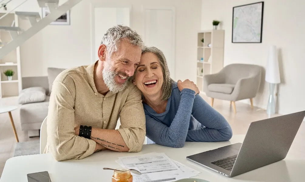 A laughing senior couple sits at a white table looking at a laptop in a bright, modern living room