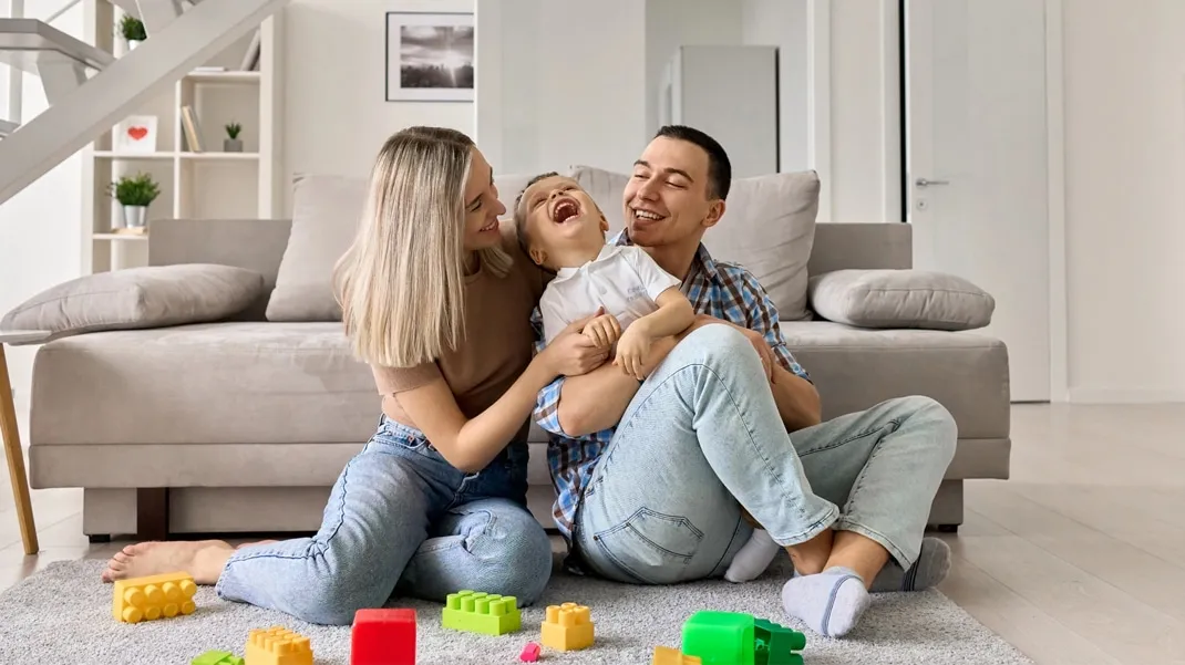 A mother and father are sitting on the floor in their living room, laughing and hugging their young child who is sitting between them.