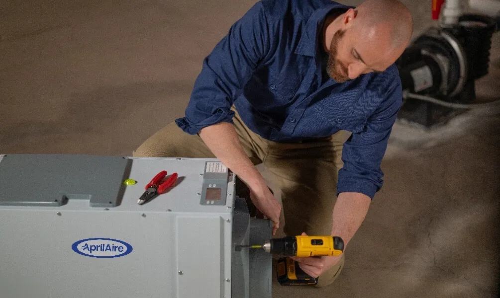 A technician uses a power drill to install an AprilAire dehumidifier unit on a concrete floor.