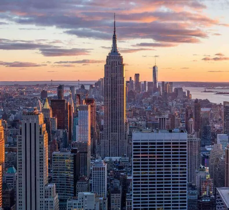The Empire State Building towers over the Manhattan skyline during a golden sunset with pink-tinged clouds.