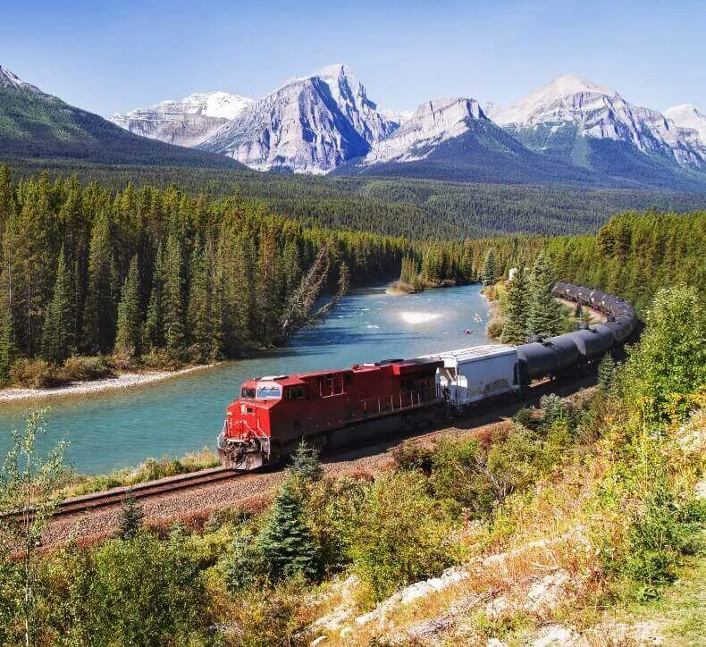 Red freight train traveling along a turquoise river and evergreen forest with snow-capped mountain peaks behind it.