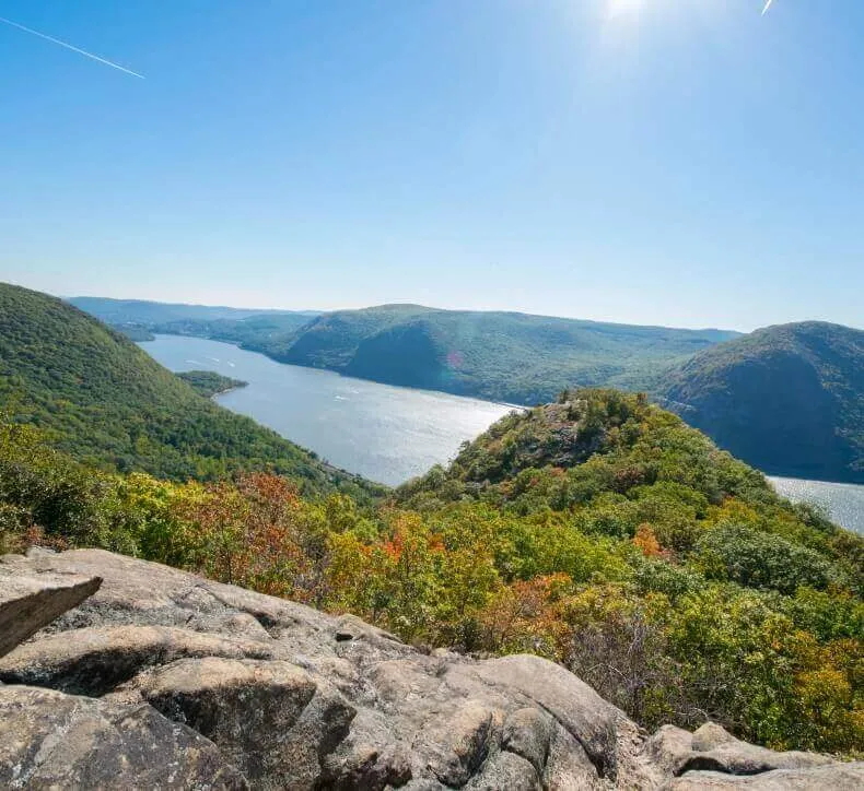 Sun shines over a wide river winding through lush green forested mountains viewed from a rocky overlook.