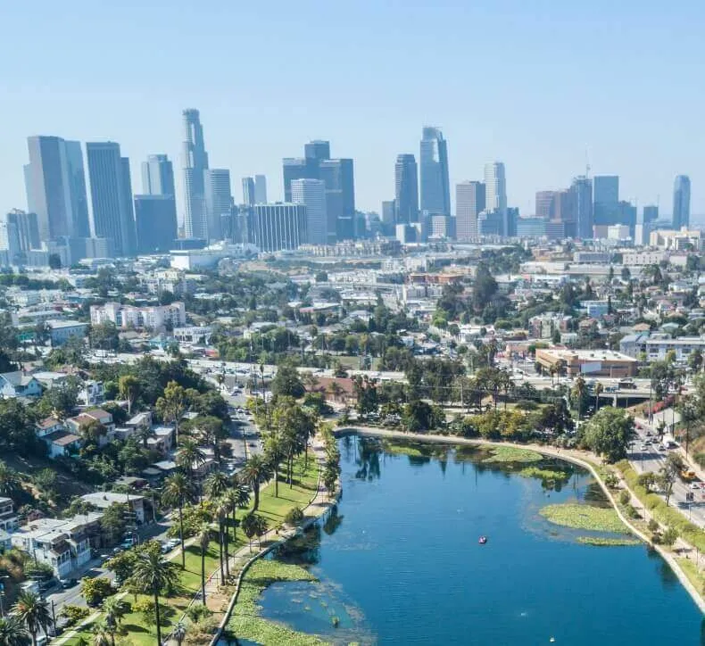 Aerial view of the Los Angeles skyline in the background, towering over a city park with a large lake and palm trees.