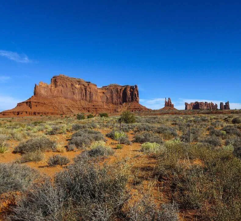 Wide view of a desert landscape featuring orange soil, scrub brush, and massive red rock buttes under a clear blue sky.
