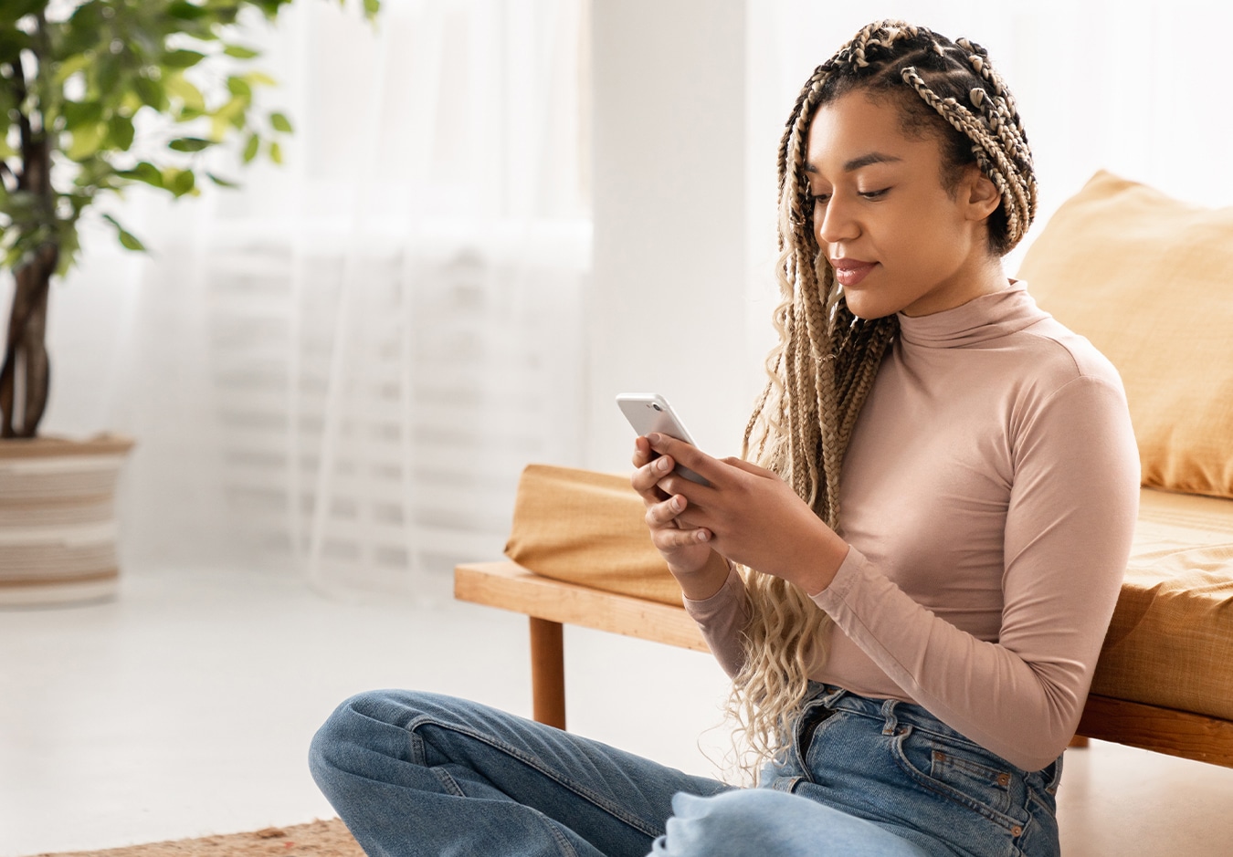 A woman with braids sits on the floor using her smartphone in a bright, modern living room.